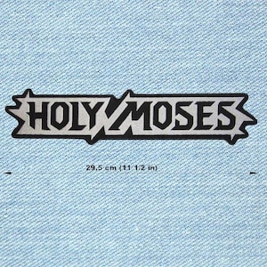 Może przedstawiać: Prostokątna naszywka z napisem "HOLY MOSES" w odważnej, stylizowanej czcionce. Tekst jest czarny z srebrnym obrysem, na tle jasnoniebieskiego dżinsu. Naszywka ma długość 29,5 cm.