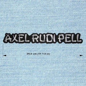 Puede incluir: Parche bordado en blanco y negro con el texto "AXEL RUDI PELL". El parche mide 29,5 cm de largo y se muestra sobre un fondo de mezclilla azul claro. El texto está en una fuente estilizada.