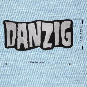 Può includere: Una toppa ricamata in bianco e nero con la scritta "DANZIG" in un carattere audace e stilizzato. La toppa misura 25 cm di larghezza e 11 cm di altezza. La toppa è su uno sfondo di denim blu chiaro.