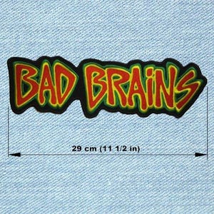 Può includere: Una toppa ricamata nera con la scritta "BAD BRAINS" in rosso e giallo. La toppa, bordata in verde e nero, misura 29 cm di lunghezza. È presentata su uno sfondo di tessuto denim azzurro chiaro.