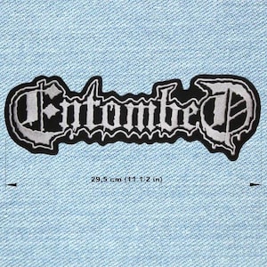 Op de afbeelding: Een zwart-wit geborduurde patch met het woord "Entombed" in een gotisch lettertype. De patch is 29,5 cm lang en heeft een golvende rand. De patch bevindt zich op een lichtblauwe denim achtergrond.
