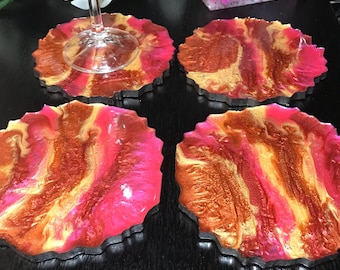 May include: Set of four pink and gold resin coasters with a marbled design. The coasters have a black edge and are perfect for protecting surfaces from heat and spills.