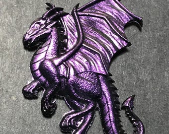 Puede incluir: Una figurita de dragón púrpura con las alas extendidas, de pie sobre una superficie gris oscuro. El dragón tiene una superficie detallada y texturizada y una cola larga.