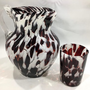 May include: A glass pitcher and a matching glass tumbler featuring a speckled pattern of deep maroon and opaque white glass. The pitcher has a clear glass handle and a wide pouring spout, while the tumbler has a cylindrical shape.