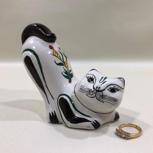 May include: A white ceramic cat figurine with black accents and floral details. The cat is in a stretching pose, with its tail arched. A gold ring with three small diamonds is next to the cat.