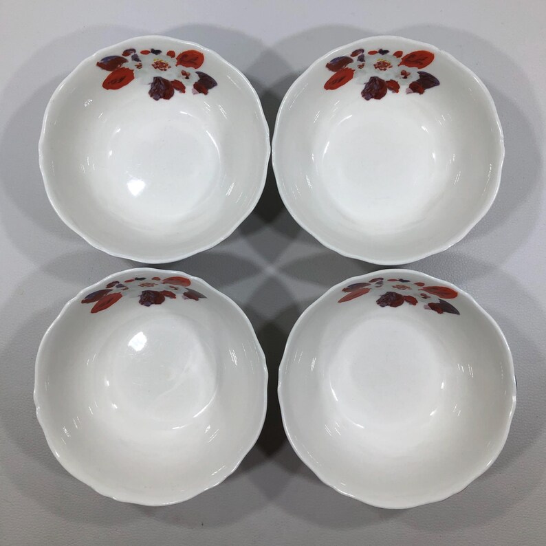 May include: Four white ceramic bowls with scalloped edges. Each bowl features a floral design in shades of red, brown, and white near the rim. The bowls are arranged in a square formation, showcasing their simple elegance.