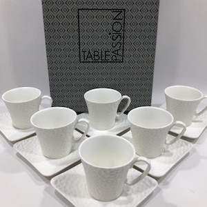 May include: A set of six white teacups and saucers with a textured surface. The cups have handles and sit on square saucers. A box with the word "TABLE" is in the background.