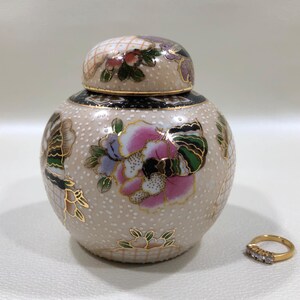 May include: A beige ceramic jar with a lid, adorned with floral and butterfly designs in pink, green, and gold. The jar has a textured surface with small white dots. A gold ring with three small diamonds is next to the jar.