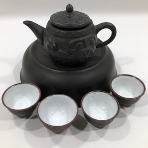 May include: A black ceramic teapot with intricate carvings sits on a matching base, surrounded by four small black teacups with white interiors.