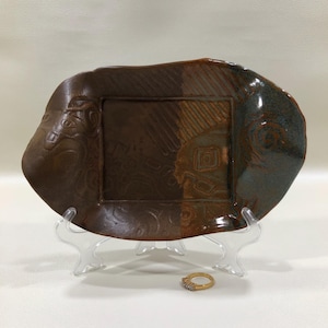May include: A handmade, rectangular ceramic plate with a brown and blue glaze. The plate has an irregular edge and a square, recessed center with etched designs. A gold ring with small stones rests in front of the plate.