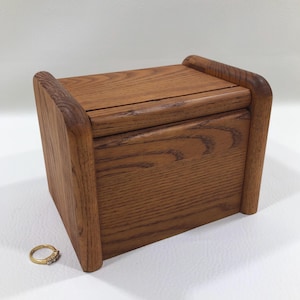 May include: A rectangular wooden box with a hinged lid and rounded side supports, featuring a prominent natural wood grain pattern in a medium brown tone. A small gold-colored ring with embedded stones is positioned next to the box.