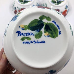 May include: White porcelain bowls with floral designs in red, blue, and green. The bottom of the bowl features the words "Porcelain", "dishwasher safe", and "made in Indonesia" in blue script. Several bowls are visible.