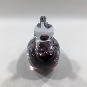 May include: A clear and deep purple glass swan figurine. The swan has a long neck and a detailed body. The glass is smooth and reflects light, creating a shimmering effect. The figurine is a decorative item.