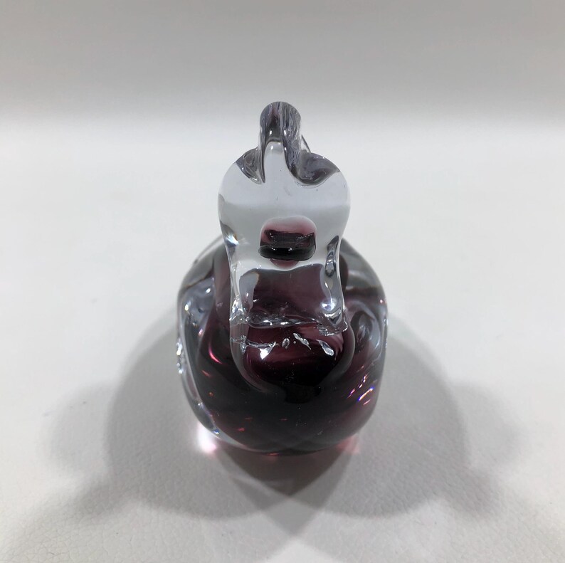 May include: A clear and deep purple glass swan figurine. The swan has a long neck and a detailed body. The glass is smooth and reflects light, creating a shimmering effect. The figurine is a decorative item.