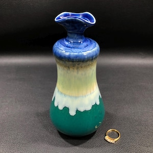 May include: A handcrafted ceramic vase with a fluted rim, showcasing a gradient of colors from deep teal to light blue. A gold ring with clear stones rests beside the vase. The vase is approximately 7 inches tall.