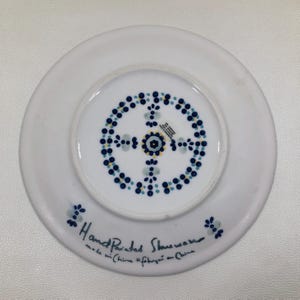 May include: White, hand-painted stoneware plate with a blue and yellow floral design on the underside. The plate has a circular pattern of blue dots and a central floral motif. Text on the plate reads "Hand Painted Stoneware made in China".