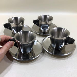 May include: A set of four stainless steel espresso cups and matching saucers. Each cup features a polished metallic finish with a black handle. The cups are tapered toward the base and sit on circular, reflective stainless steel saucers.