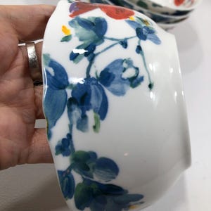 May include: White ceramic bowl with a floral design featuring blue and red flowers and green leaves. The bowl is held in a hand, with other bowls visible in the background. The floral pattern is hand-painted.