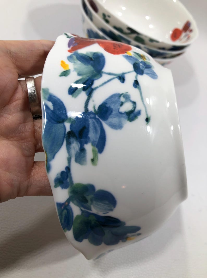 May include: White ceramic bowl with a floral design featuring blue and red flowers and green leaves. The bowl is held in a hand, with other bowls visible in the background. The floral pattern is hand-painted.