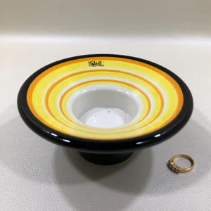 May include: A ceramic bowl with a black base and rim, featuring concentric yellow and orange stripes. The interior is white. A gold ring with small stones sits to the side. The bowl has the word "Faldelli" printed on it.
