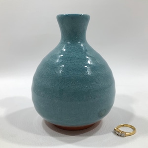 May include: A turquoise ceramic vase with a crackle glaze finish. The vase has a rounded body and a narrow neck. A gold ring with three small diamonds sits next to the vase.