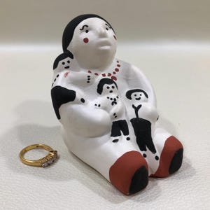 May include: A white ceramic figurine of a seated figure holding three smaller figures. The figurine has black hair, red accents, and a red necklace. A gold ring with three stones sits in front of the figurine.