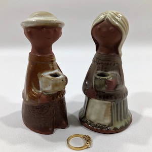 May include: A pair of ceramic candle holders in the shape of a man and woman. The man is wearing a brown hat and a brown coat. The woman is wearing a brown sweater and a white skirt. Both are holding a small cup in their hands.