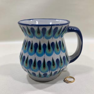 May include: A white ceramic mug with a rounded body and a blue handle, featuring a repeating pattern of blue, teal, and black teardrop shapes. A small gold-toned ring with clear stones is positioned on the surface next to the base of the mug.