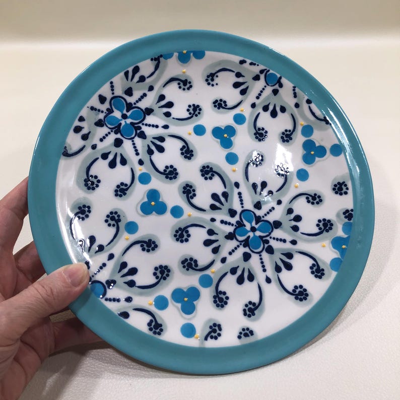May include: A round ceramic plate with a turquoise rim and a white center. The plate features a repeating floral pattern in shades of blue and small yellow accents. The plate is held by a hand.