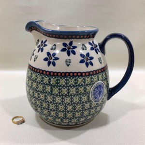 May include: A ceramic pitcher with a blue handle and rim, decorated with blue floral and green leaf patterns. The pitcher has a bulbous shape and a spout. A gold ring with small stones sits next to the pitcher.