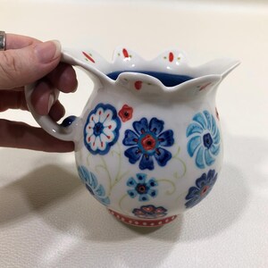 Puede incluir: Jarra de cer&aacute;mica blanca con borde festoneado y asa. La jarra est&aacute; decorada con motivos florales azules y rojos. El interior es de color azul oscuro. La base tiene un borde rojo punteado.