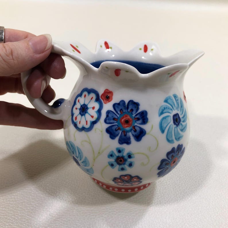 Puede incluir: Jarra de cer&aacute;mica blanca con borde festoneado y asa. La jarra est&aacute; decorada con motivos florales azules y rojos. El interior es de color azul oscuro. La base tiene un borde rojo punteado.