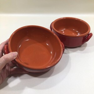 May include: Two red ceramic bowls with handles. The bowls have a round shape and a deep orange interior. The exterior is a darker red. The bowls are likely for serving soup or other dishes.