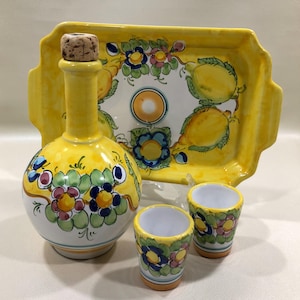 May include: A yellow ceramic liquor bottle with a cork stopper, a serving tray, and two shot glasses. The set is decorated with floral and lemon designs. The bottle and shot glasses have a white base. The tray has handles.