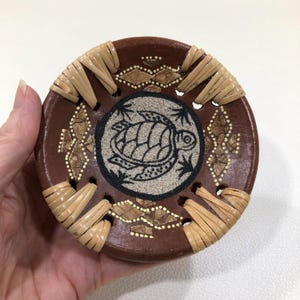 May include: A round, brown decorative plate with a turtle design in the center. The plate has woven straw accents around the edge and decorative patterns. The turtle is black and the background is tan.