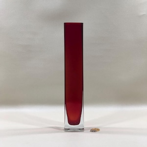 May include: A tall, rectangular, ruby-red glass vase. The vase has a clear, rectangular base and a smooth, polished surface. The vase is empty and stands upright on a white surface. A small, gold ring is next to the vase.