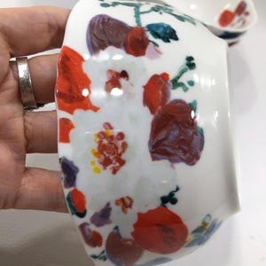 May include: White ceramic bowl with a floral design. The bowl features a hand-painted pattern of red, purple, and white flowers with green leaves. The bowl is oval-shaped and has a glossy finish. Additional bowls are visible in the background.