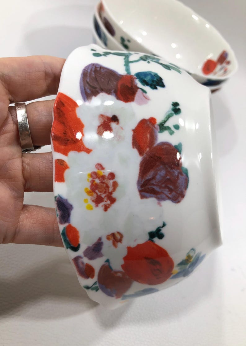 May include: White ceramic bowl with a floral design. The bowl features a hand-painted pattern of red, purple, and white flowers with green leaves. The bowl is oval-shaped and has a glossy finish. Additional bowls are visible in the background.