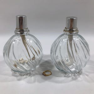 May include: Two clear glass oil lamps with a spiral design and silver-colored metal tops. A gold ring with three small stones sits in front of the lamps. The lamps have wicks extending from the top.