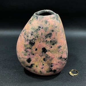 May include: A handcrafted ceramic vase with a unique, organic shape. The vase features a blend of pink, black, and yellow hues, creating a textured, bubble-like pattern. A small gold ring with three small stones sits nearby. The vase has an opening at the top.