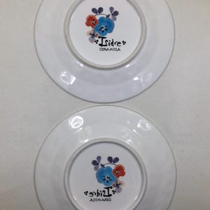 May include: Two white ceramic plates with a floral design in the center. The design includes blue, red, and purple flowers. The text "Isidre CERAMICA" is printed on the plates.