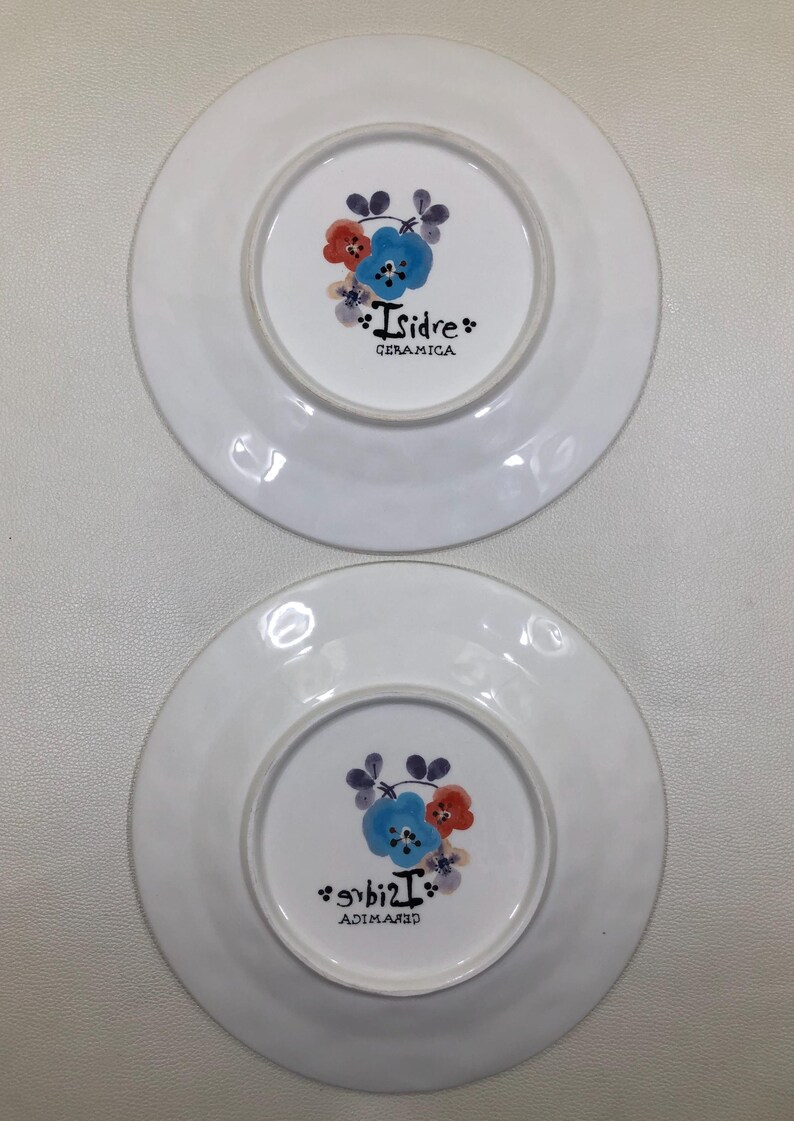 May include: Two white ceramic plates with a floral design in the center. The design includes blue, red, and purple flowers. The text "Isidre CERAMICA" is printed on the plates.