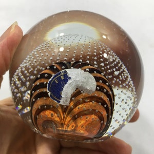 May include: A clear glass paperweight with a swirling pattern of brown and black lines inside. The paperweight is filled with small air bubbles and has a blue sticker with the text "ER" on it.