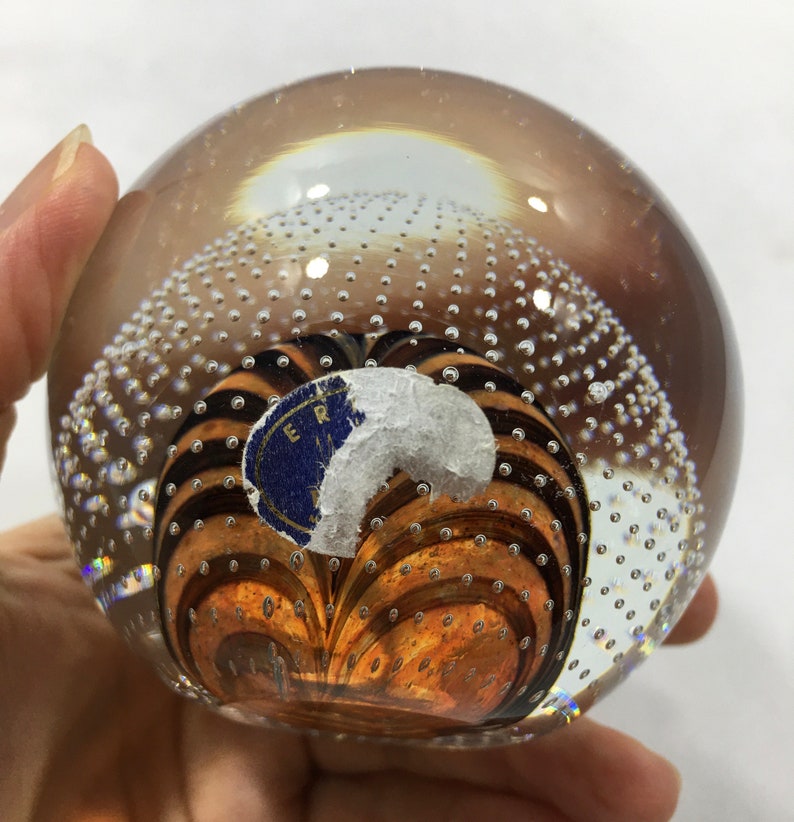 May include: A clear glass paperweight with a swirling pattern of brown and black lines inside. The paperweight is filled with small air bubbles and has a blue sticker with the text "ER" on it.