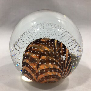 May include: A clear glass paperweight with a brown and white swirl pattern inside. The paperweight is filled with small air bubbles.
