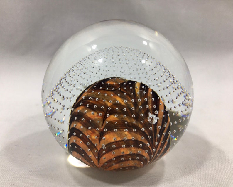 May include: A clear glass paperweight with a brown and white swirl pattern inside. The paperweight is filled with small air bubbles.