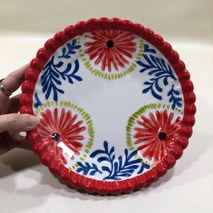 May include: A round, white ceramic plate with a scalloped red rim. The plate features a floral design with red, blue, and green accents. The center of the plate has a white background. The plate is approximately 20 cm in diameter.