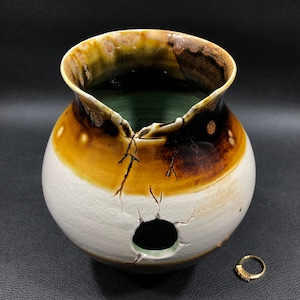 May include: A white ceramic vase with brown and green glaze. The vase has a wide mouth and a round body. There is a hole in the side of the vase.