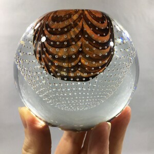 May include: A clear glass paperweight with a brown and white swirl design. The paperweight is filled with small air bubbles.