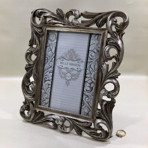 May include: Ornate silver-colored picture frame with scrolling floral details. The frame holds a 5x7 inch print with a floral design and the text "BELLE MAISON". A small gold ring with a diamond is next to the frame.